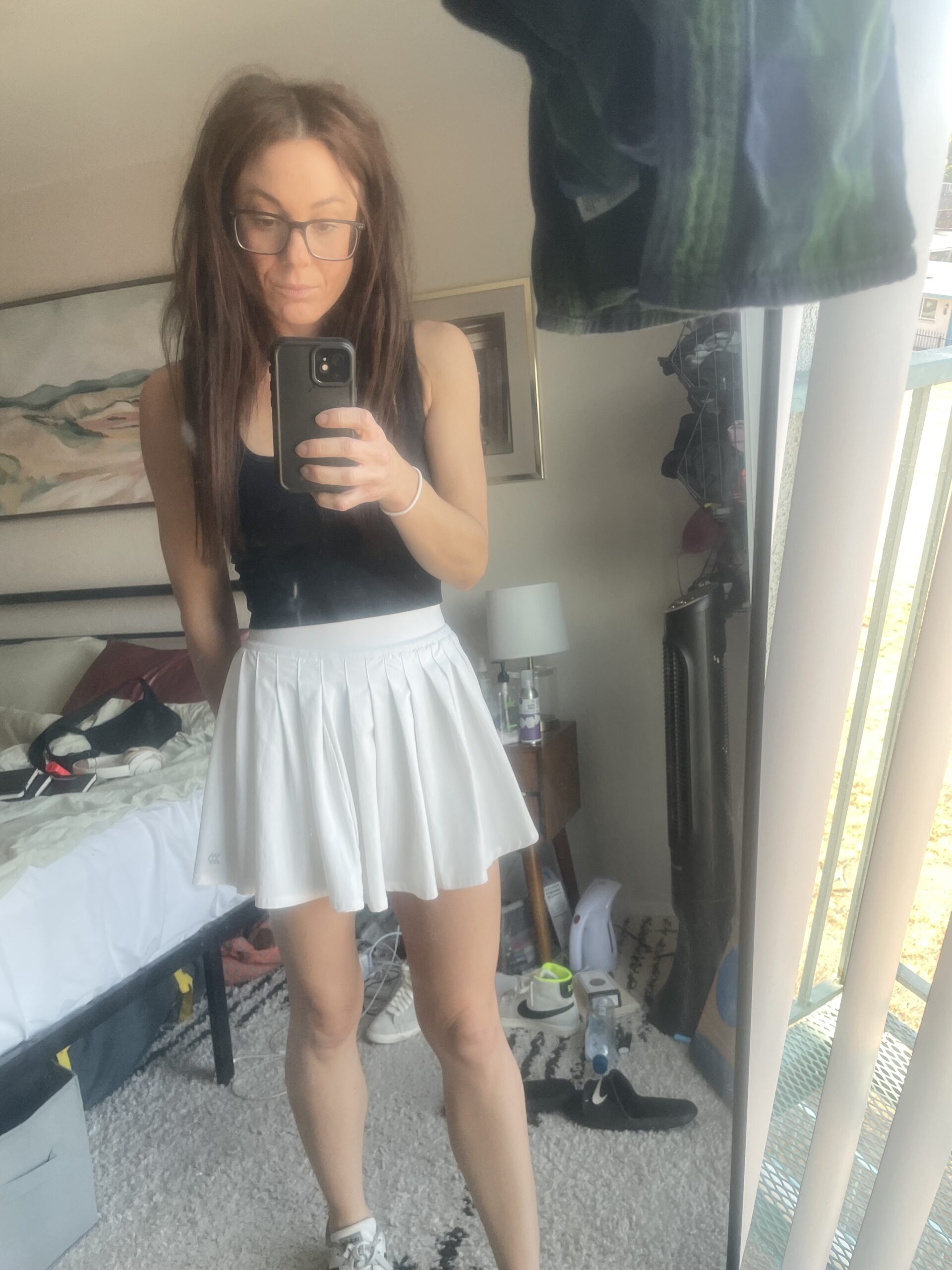 Woman wearing a black tank top and a white pleated athletic skirt from Target's JoyLab collection takes a mirror selfie in a bedroom. The background includes a bed, a nightstand, and various items scattered around the room.