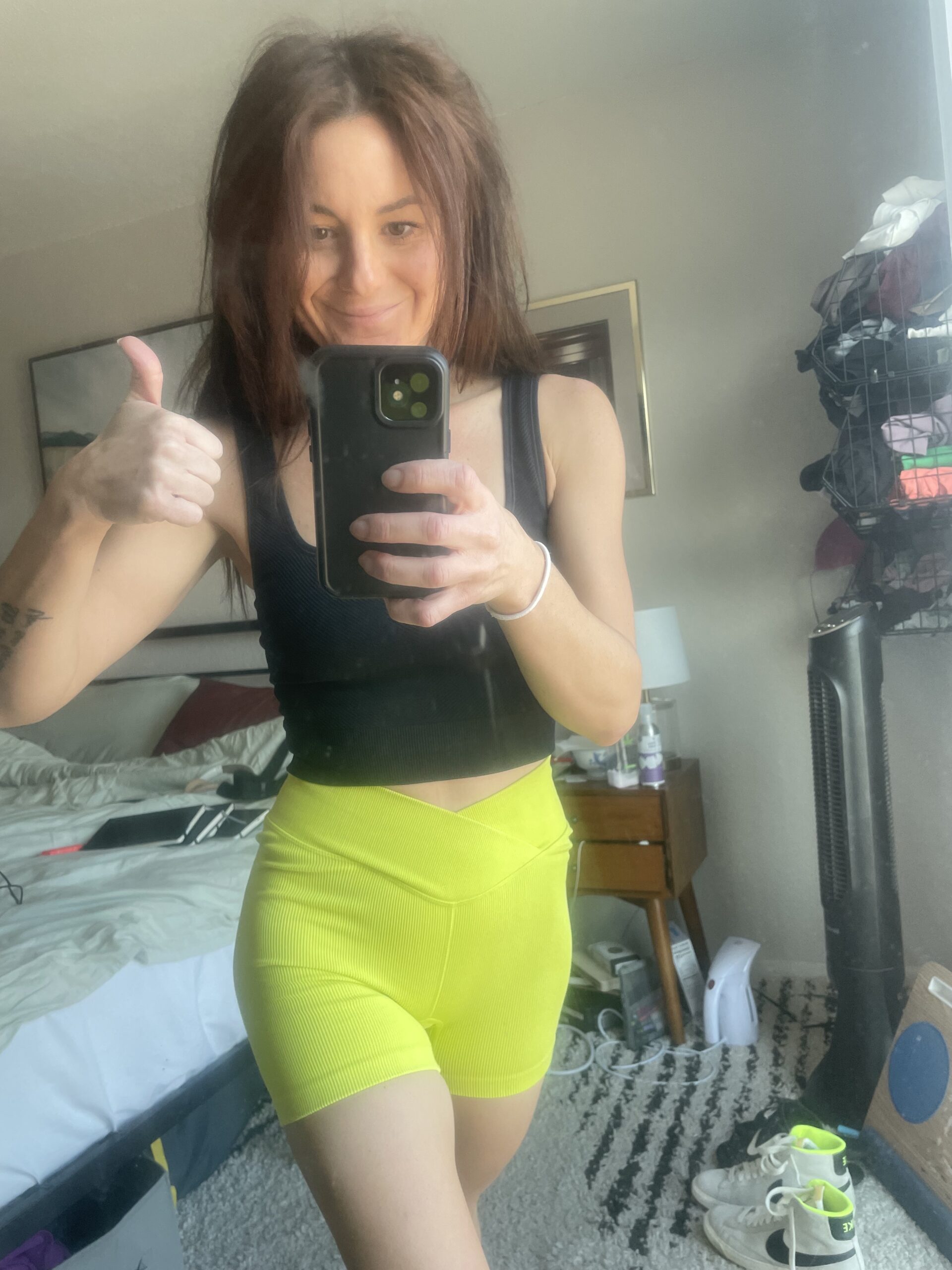 Woman wearing a black tank top and neon yellow high-waisted athletic shorts from Target's All in Motion collection gives a thumbs up while taking a mirror selfie in a bedroom. The background features a bed, nightstand, and a fan.