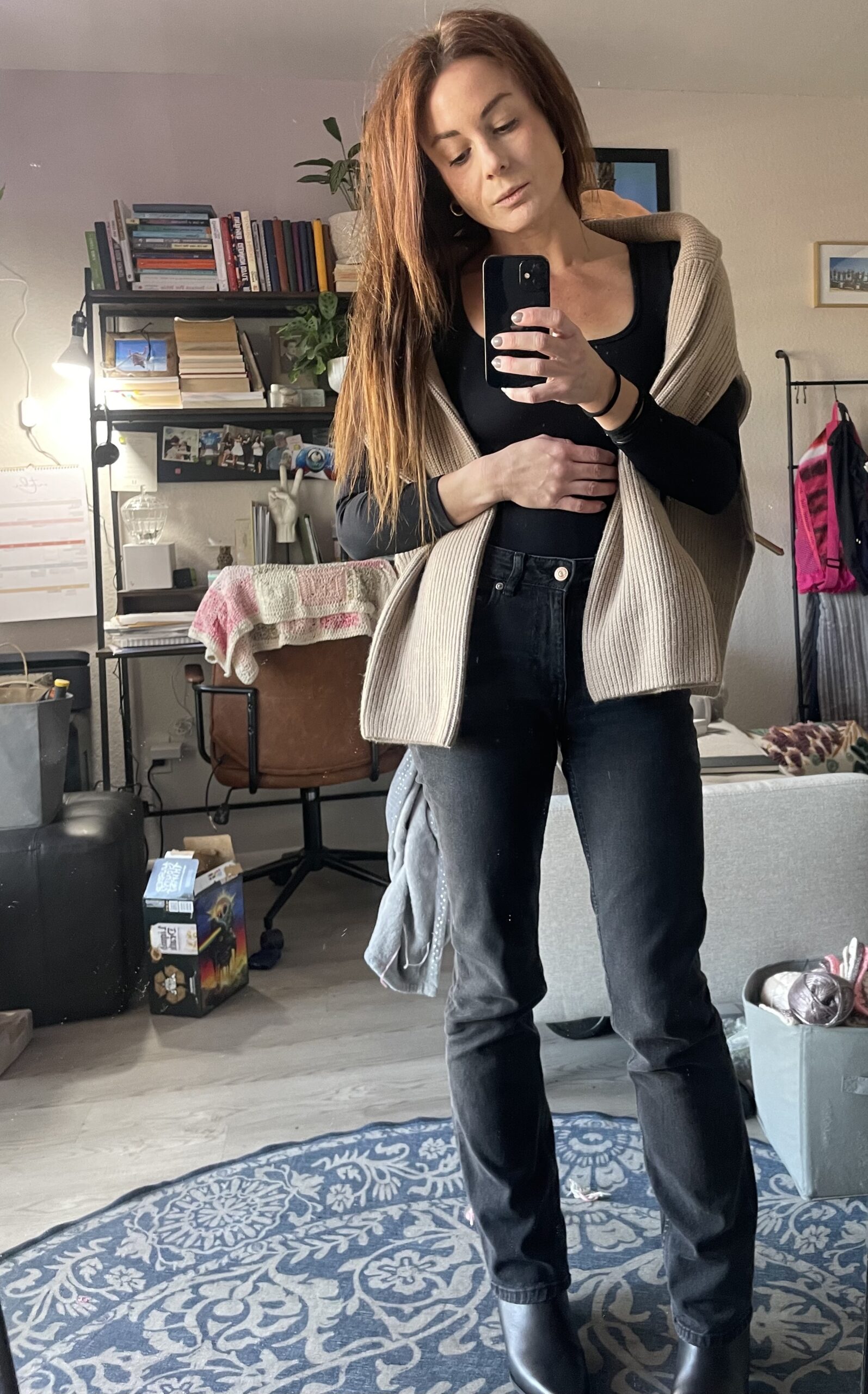 Woman wearing Amazon black boots, black jeans, and a black top with a beige cardigan draped over her shoulders, standing in a cozy, well-decorated room.
