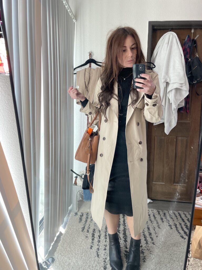 Woman wearing black boots, a black dress, and a beige trench coat, holding a tan handbag, posing in front of a mirror in a bright room.