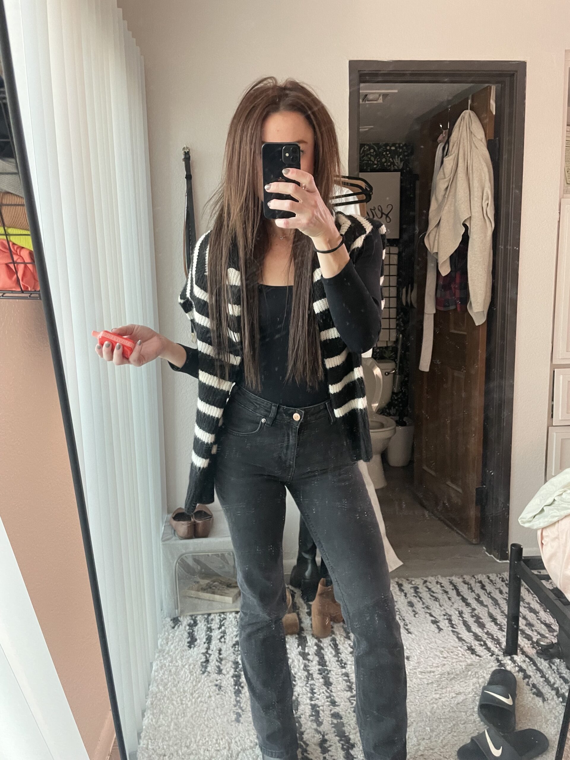 Woman in a black outfit with a striped black and white cardigan takes a mirror selfie. She stands in a room with various shoes and clothes in the background. This image is for a personal blog discussing style, mental health, exercise, habits, routines, and journaling.