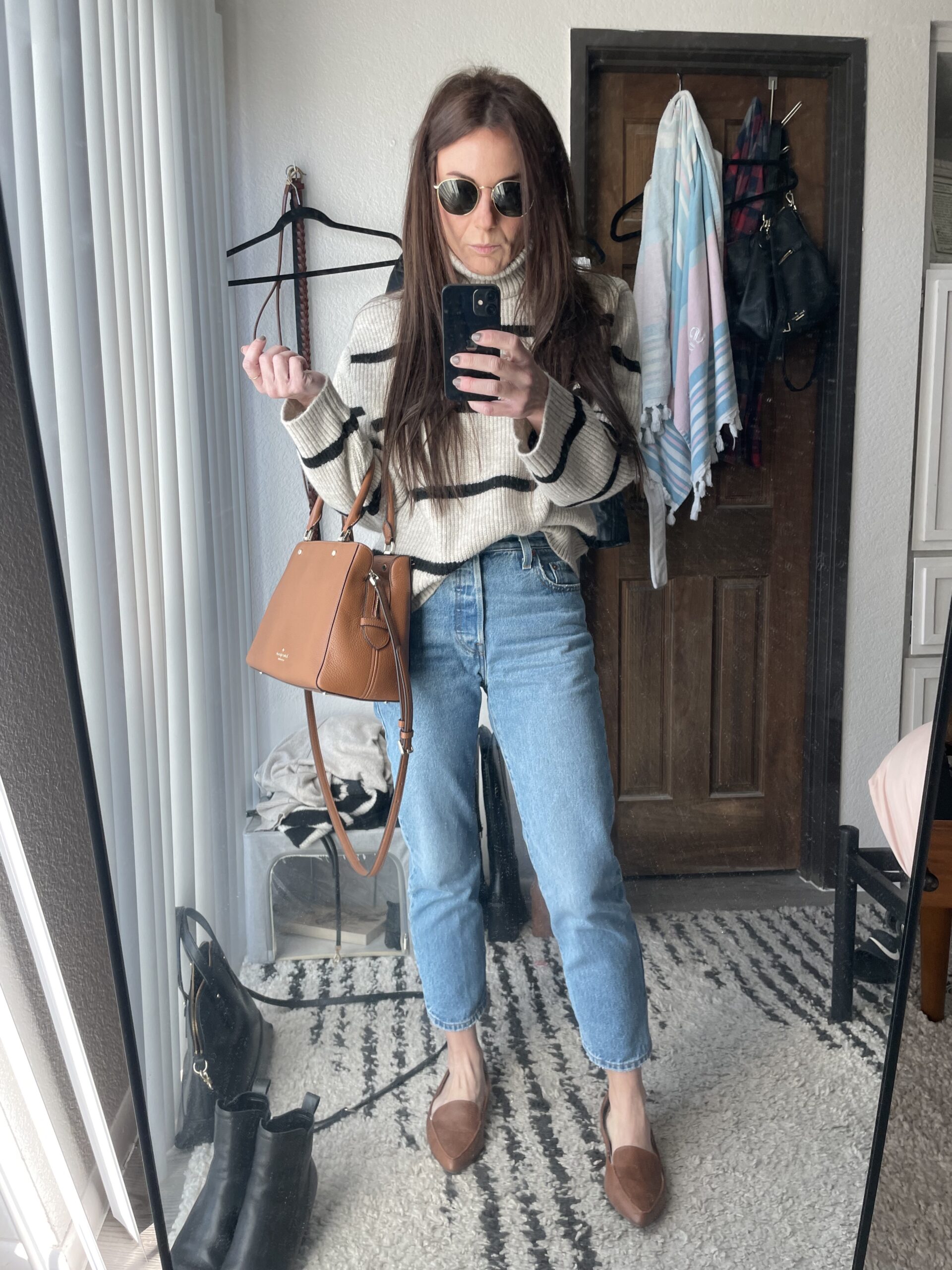 Woman wearing sunglasses, a striped sweater, and blue jeans takes a mirror selfie. She holds a brown handbag and stands in a room with clothes and accessories in the background. This image is for a personal blog discussing style, mental health, exercise, habits, routines, and journaling.
