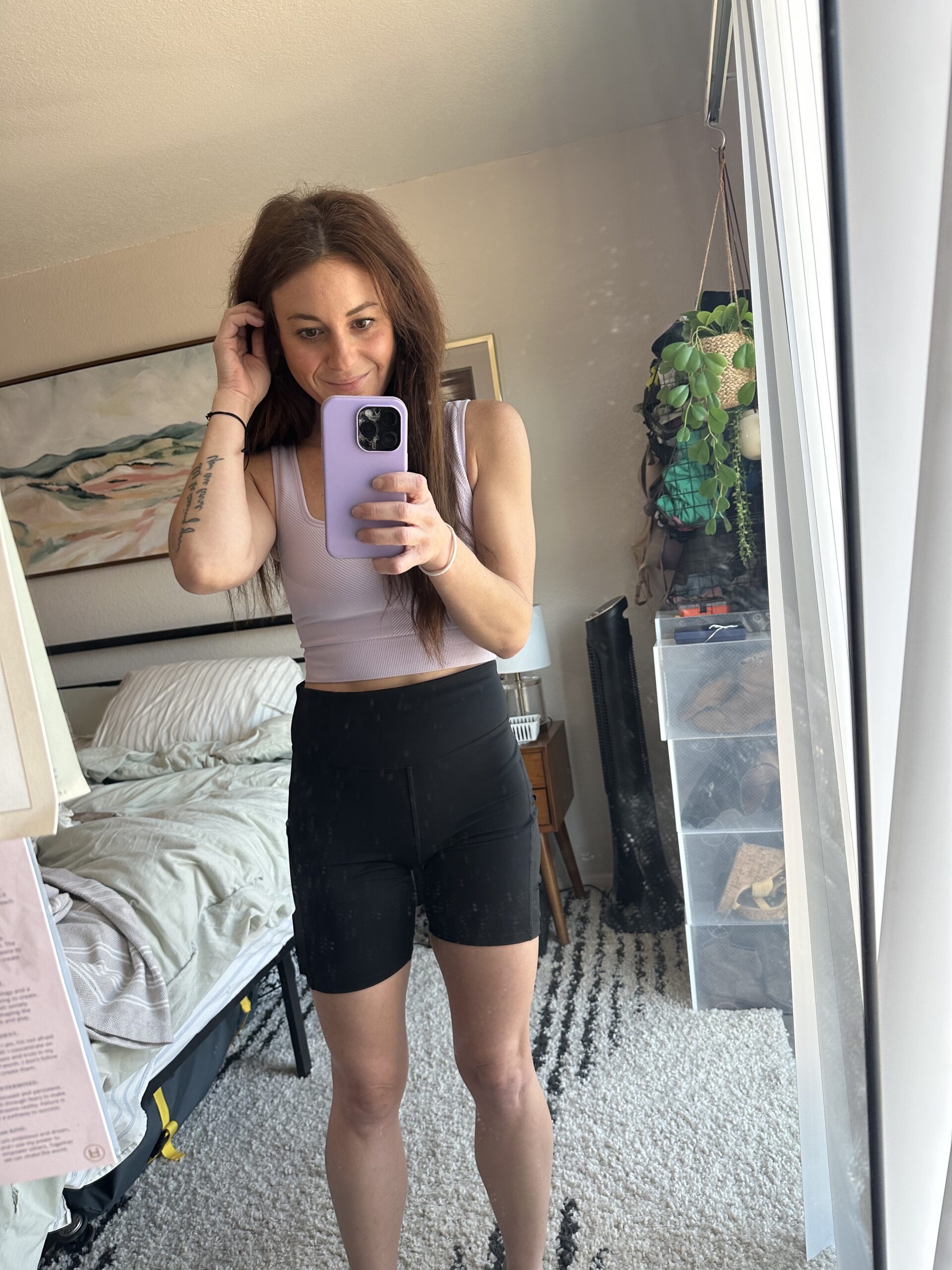 Woman in a light purple tank top and black high-waisted athletic shorts from Target's All in Motion collection takes a mirror selfie in a bedroom. The background includes a bed, nightstand, and various personal items.