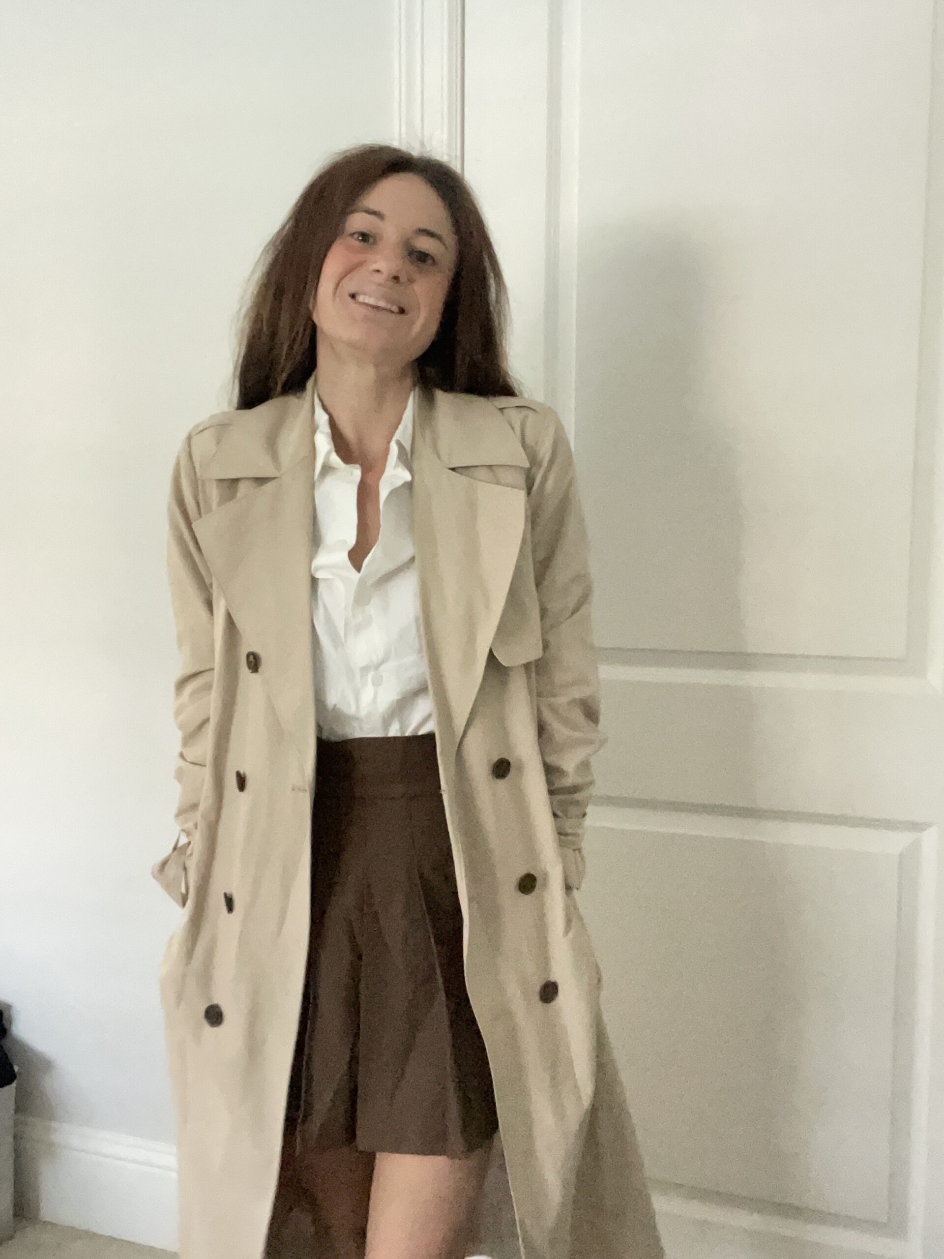 Woman standing indoors wearing a beige trench coat over a white button-down shirt and a brown skirt, smiling at the camera.