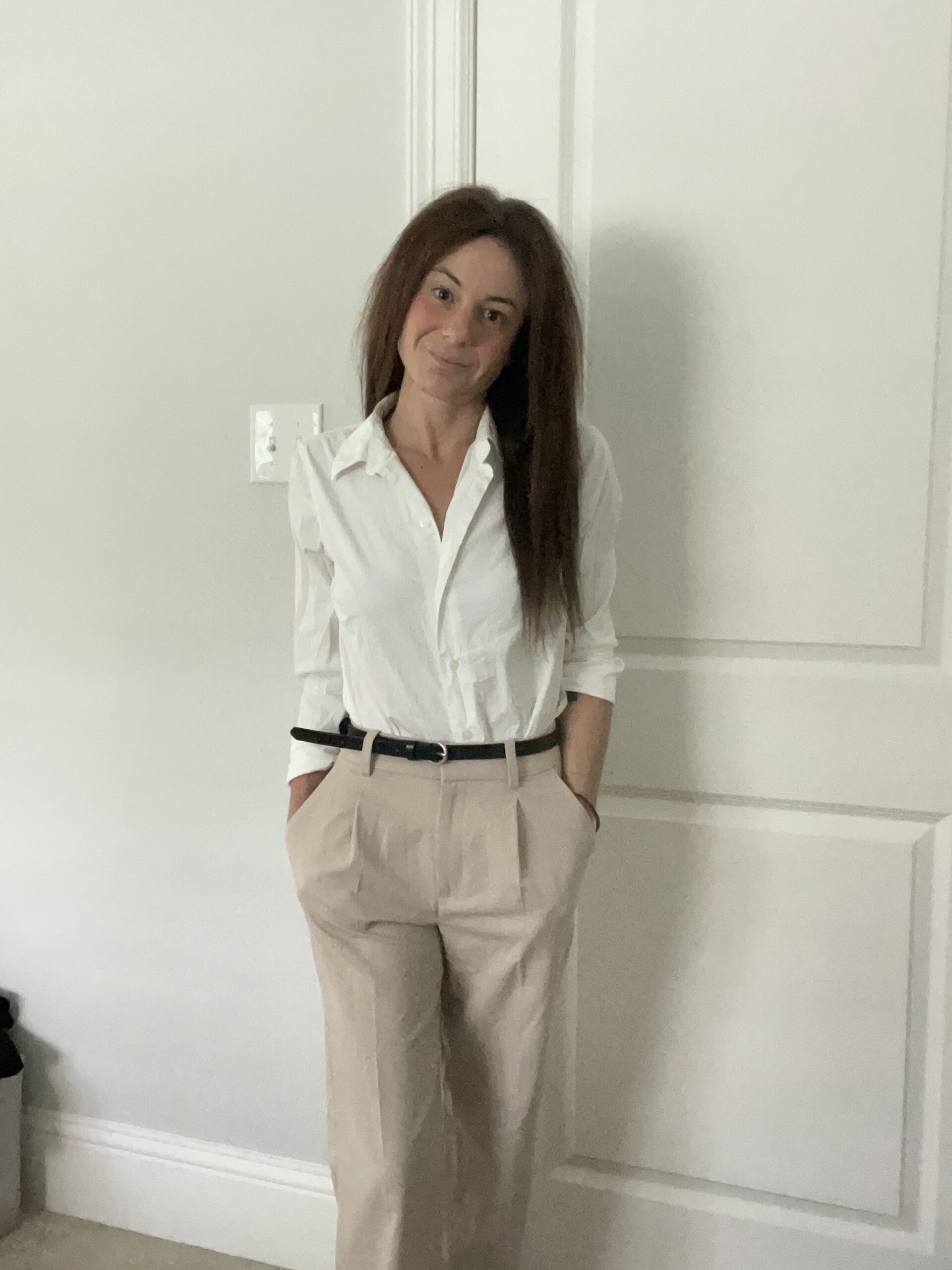 Woman standing indoors wearing a white button-down shirt tucked into beige high-waisted trousers, with hands in pockets and smiling.