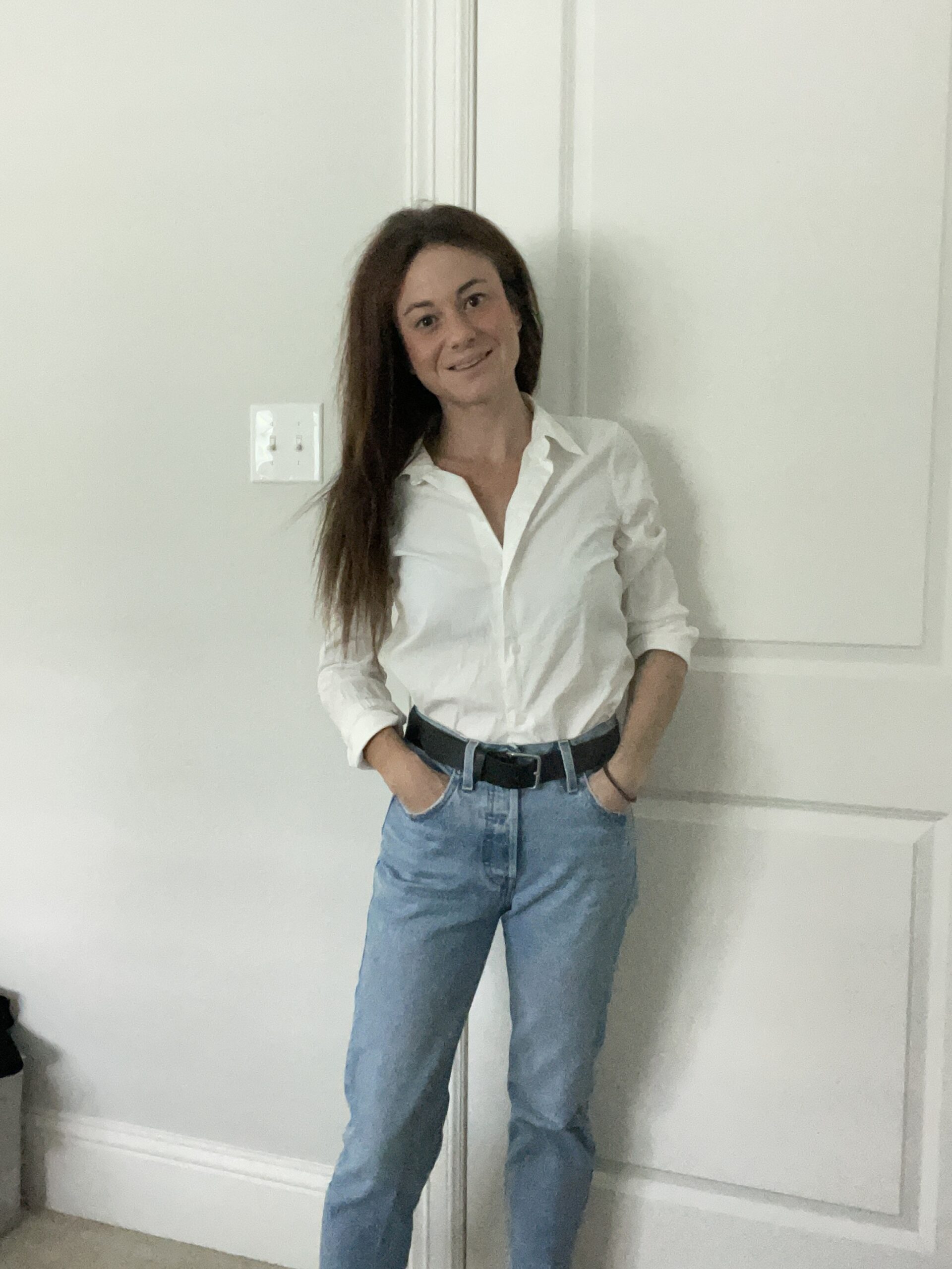 Woman standing indoors wearing a white button-down shirt tucked into blue jeans, with hands in pockets and smiling.
