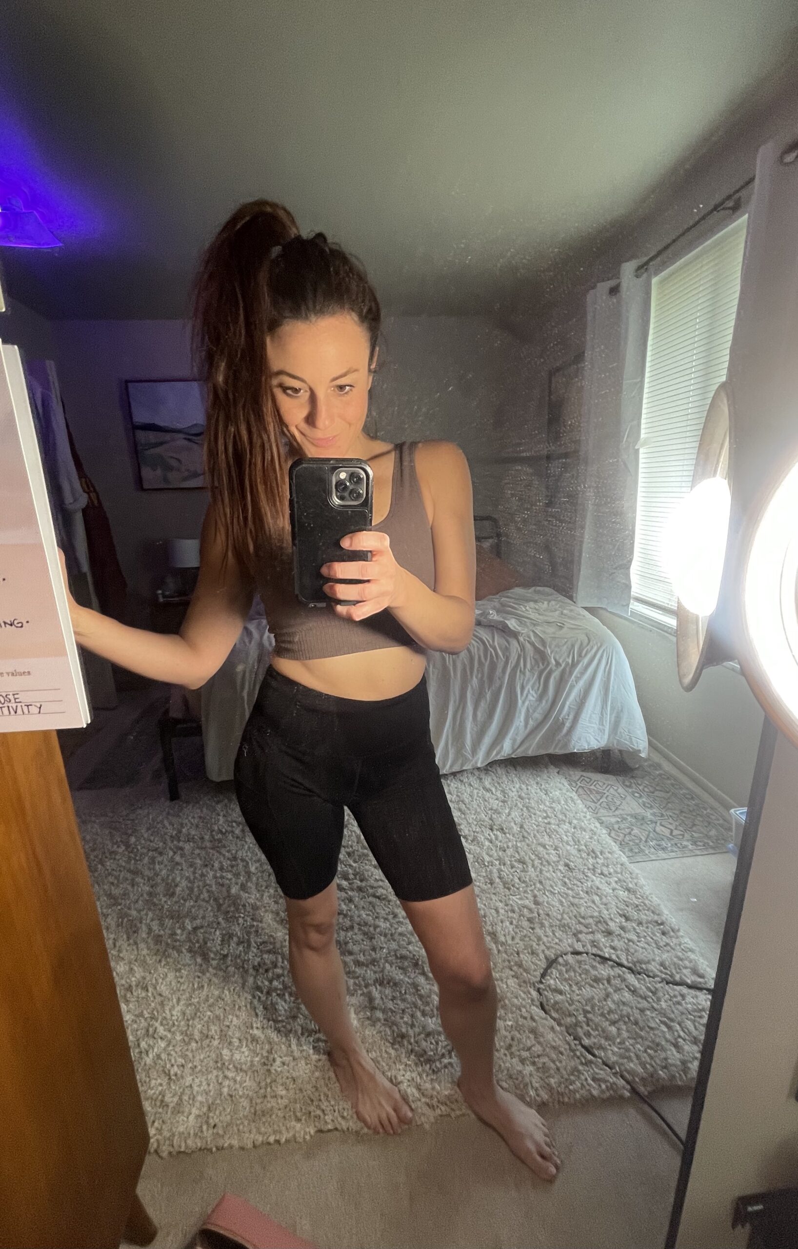 Woman wearing a gray crop top and black high-waisted bike shorts from Target's All in Motion collection takes a mirror selfie. The background features a bed, window, and various items around the room.