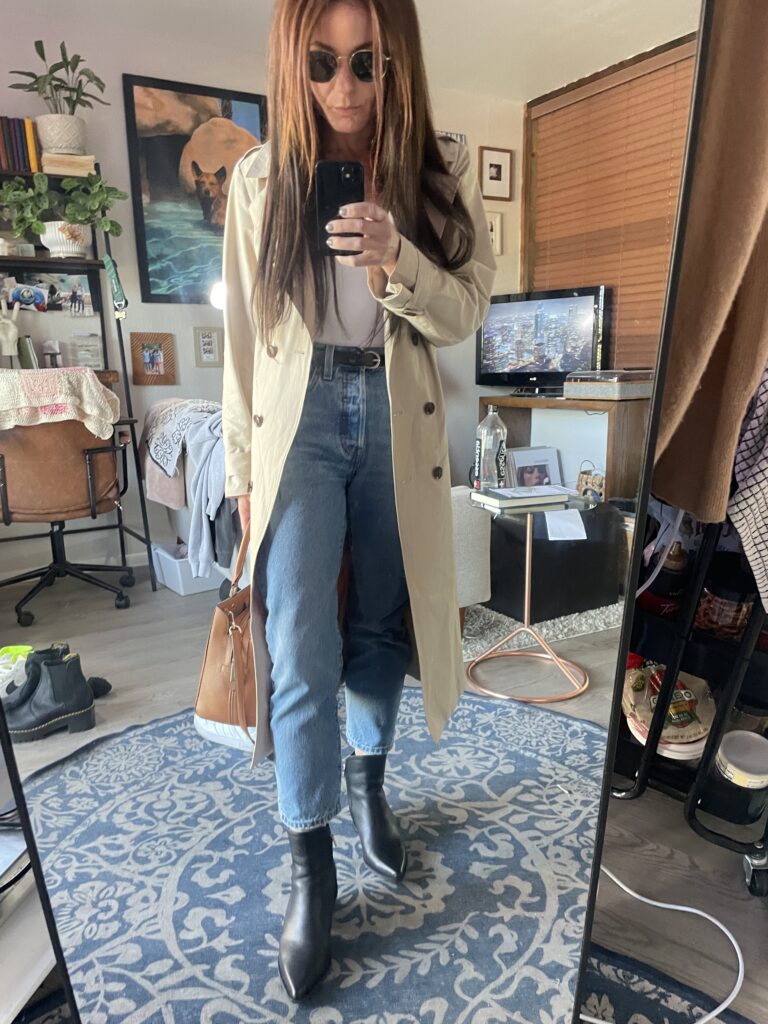 Woman wearing a beige Amazon trench coat over a white tee and light-wash jeans, paired with black boots and a brown handbag, taking a mirror selfie.