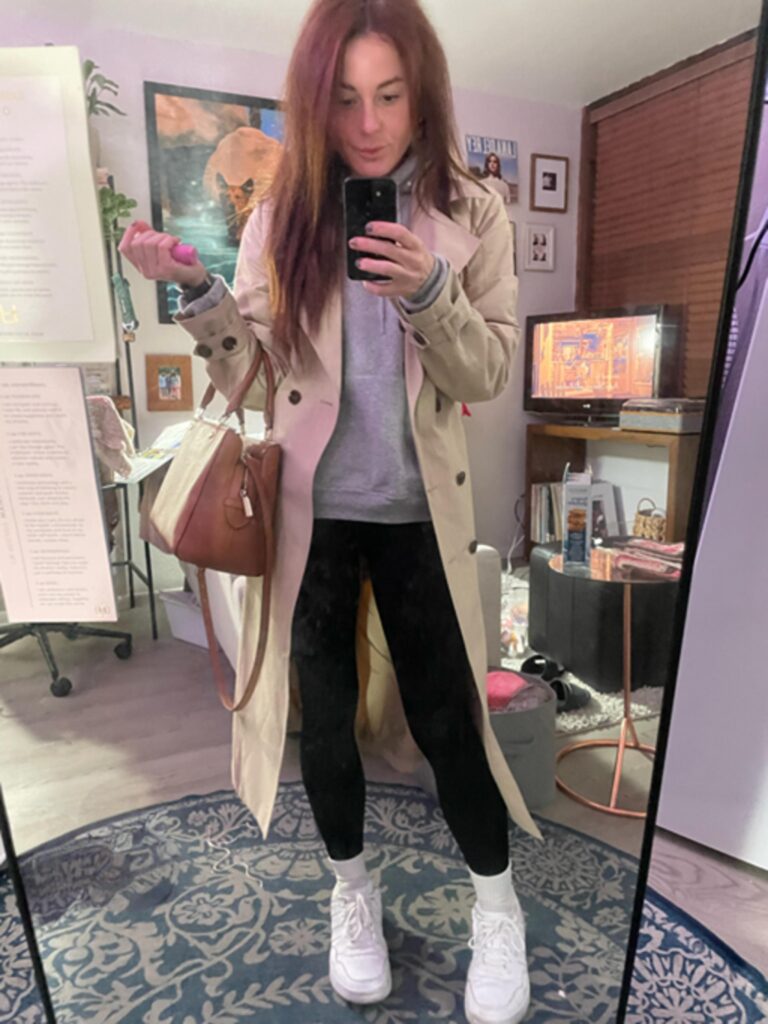 Woman in a beige Amazon trench coat over a grey hoodie and black leggings, paired with white sneakers and a brown handbag, taking a mirror selfie.