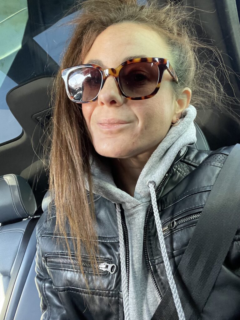 Woman wearing a black leather jacket with a grey hoodie, styled with oversized tortoiseshell sunglasses, seated in a car.