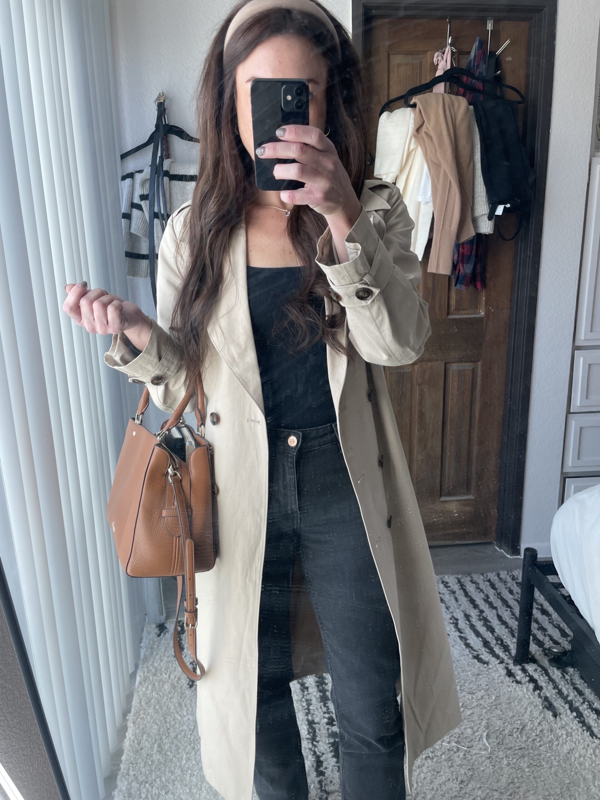 Woman wearing a beige Amazon trench coat over a black top and black jeans, accessorized with a brown handbag and a beige headband, taking a mirror selfie.