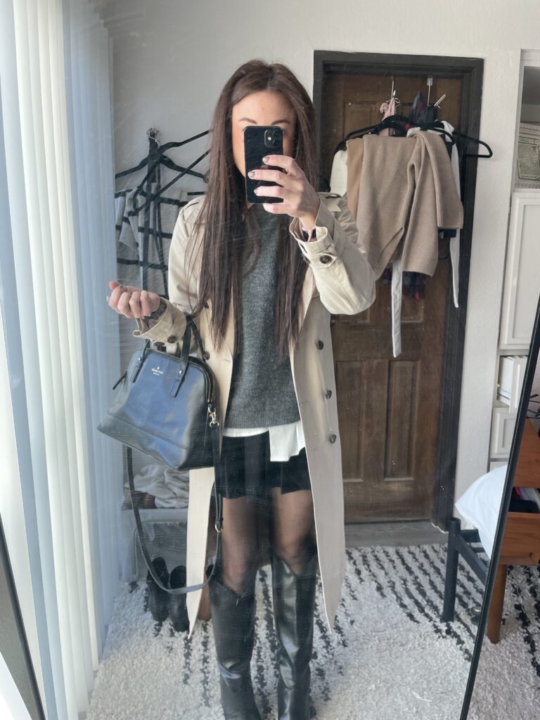 Woman in a beige Amazon trench coat over a grey sweater and black skirt, paired with black knee-high boots and a black handbag, taking a mirror selfie.