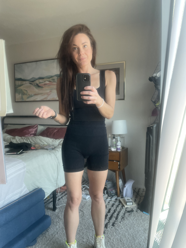 Woman taking a mirror selfie in a bedroom, wearing a black sleeveless top and black shorts, with a neutral expression and holding a phone.