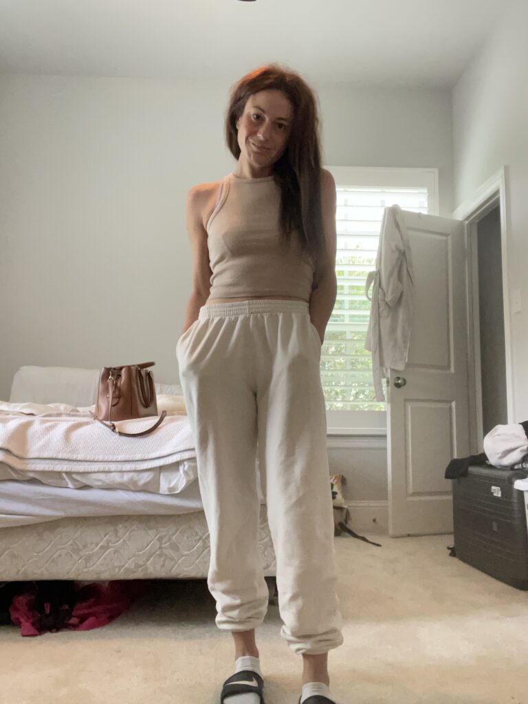 Woman standing in a bedroom wearing a beige tank top and white jogger pants, with hands in pockets, and a casual, confident expression.