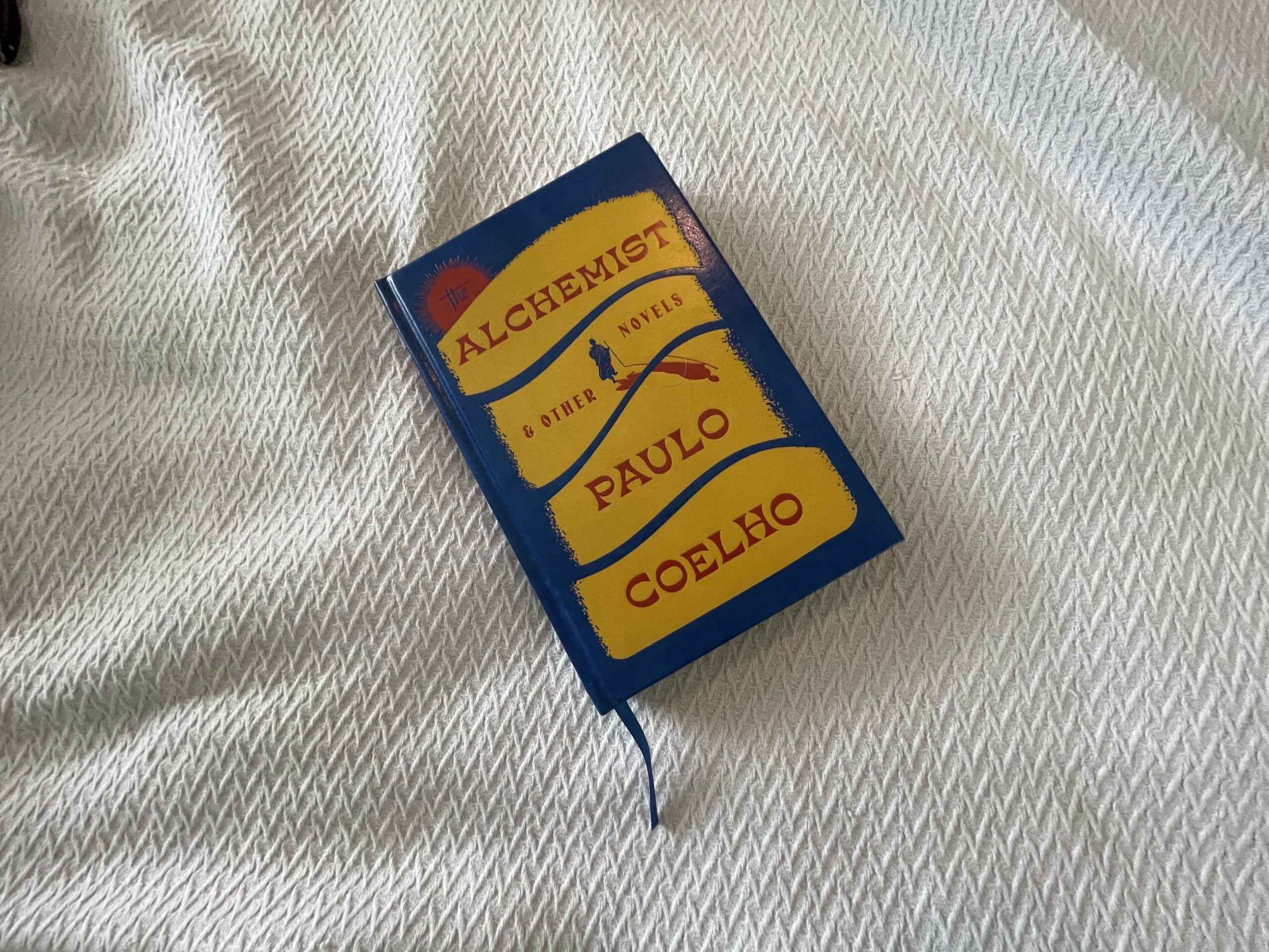 A hardcover edition of "The Alchemist" by Paulo Coelho is resting on a textured white bedspread. The book cover is predominantly blue with yellow and red accents, featuring the title and author's name prominently.