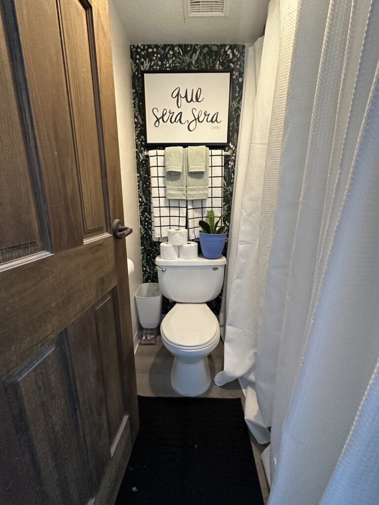 A compact bathroom with a white toilet against a dark floral-patterned wallpaper. Above the toilet is a sign that reads "que sera, sera" in cursive, with a towel rack below holding green and white towels. On the toilet tank are several rolls of toilet paper and a blue plant pot with a green plant. The bathroom door is partially open, and a white shower curtain is visible to the right.