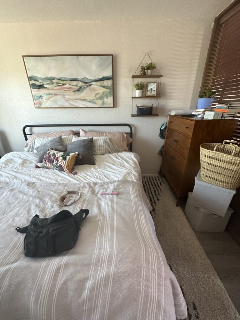A cozy bedroom with a neatly made bed covered with a light-colored, striped duvet. Above the bed is a large framed painting of a serene landscape. The bed has several pillows and a decorative blanket. To the right of the bed is a wooden dresser with various items on top, and to the left, a wall with shelves holding small potted plants and framed pictures. A black bag and a pair of headphones are placed on the bed.
