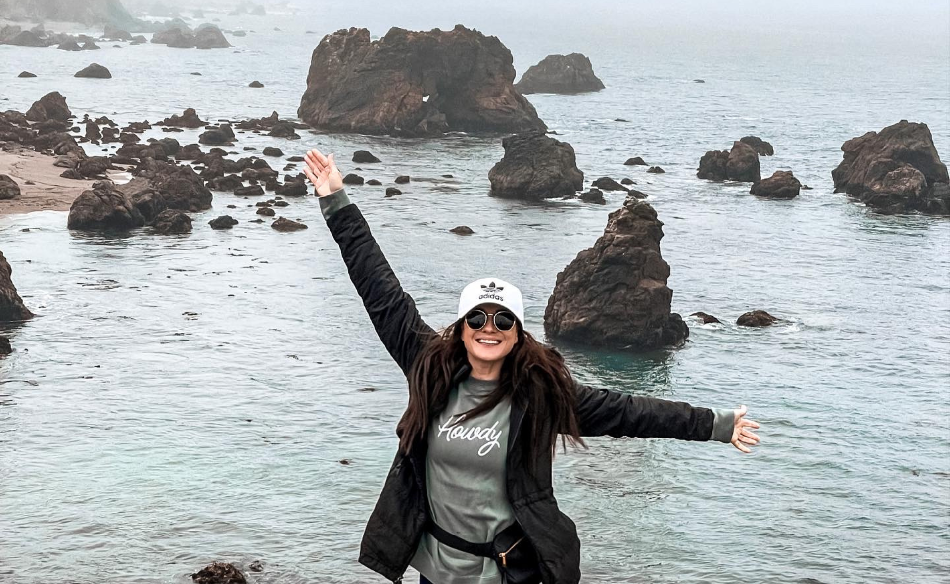 Me smiling and raising my arms in excitement while standing on a rocky coastline in California during a trip to the Redwood Forest in May 2022. This was right before I found out I was living with cPTSD.