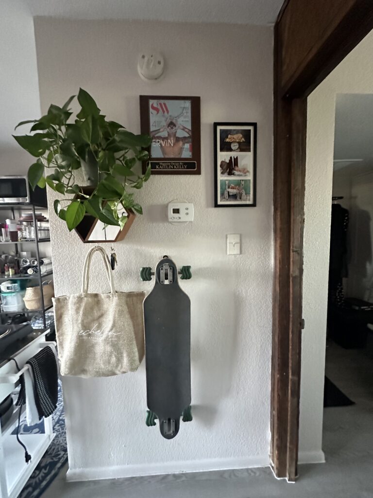 An organized entryway with a skateboard mounted vertically on the wall. Above the skateboard are a framed magazine cover, a wooden plaque, and a small framed picture. To the left, a hexagonal shelf holds a potted green plant, and a beige tote bag hangs from a hook. The area is adjacent to an open kitchen with various utensils and items visible.