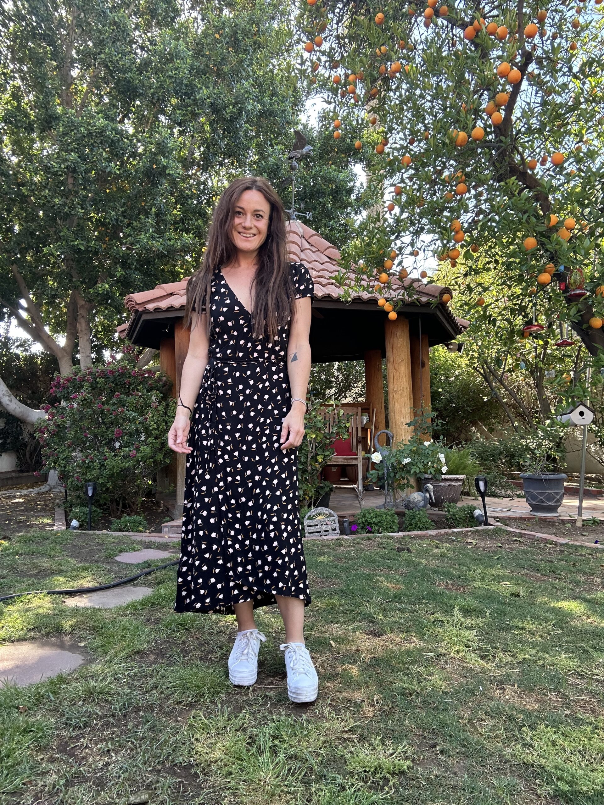 Moving into my own apartment was an exciting day. A woman with long, wavy hair is standing in a lush garden, smiling at the camera. She is wearing a black dress with a white pattern and white sneakers. Behind her is a wooden gazebo with a red-tiled roof, surrounded by various plants and trees, including an orange tree with ripe oranges hanging from its branches. The ground is a mix of grass and stone pathways.