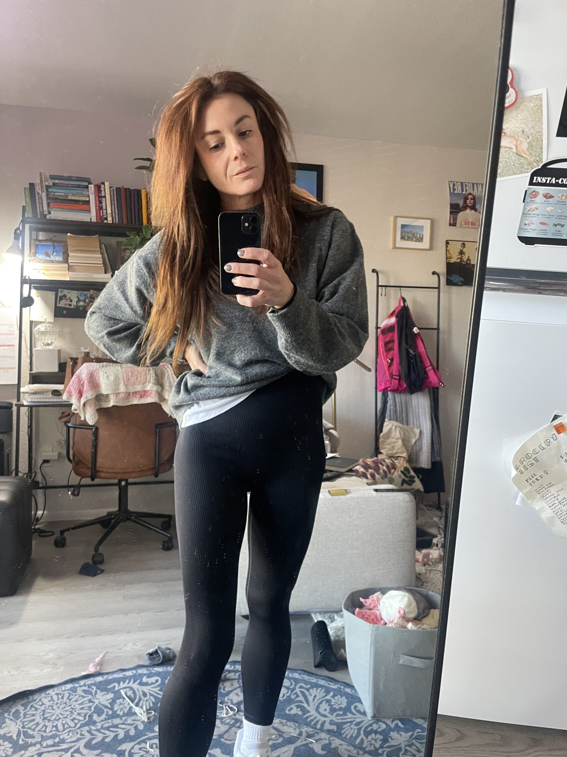 Woman taking a mirror selfie in a living room, wearing a gray sweater and black leggings, with a relaxed expression and hand on hip.